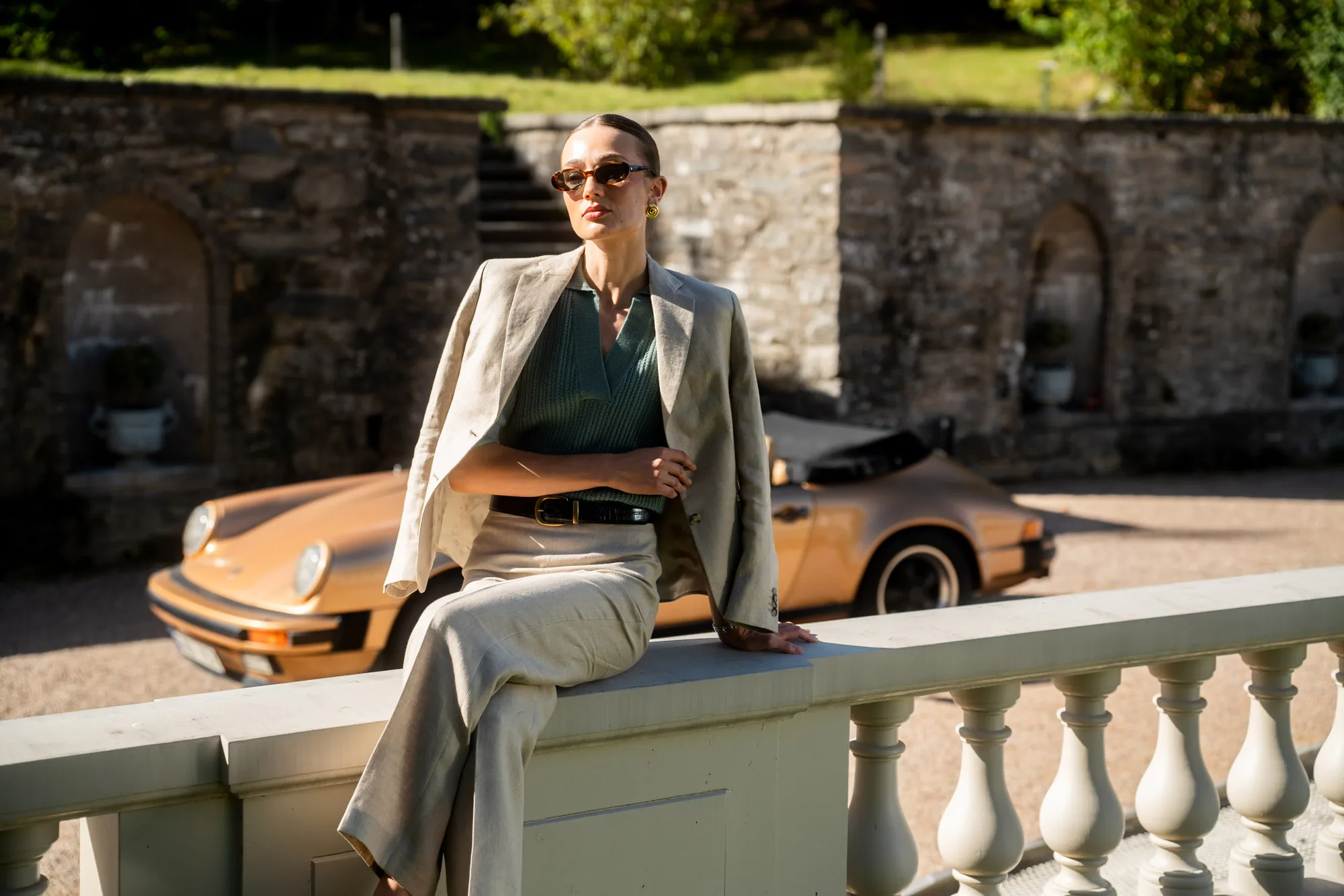 Cavaliere spring/summer campaign shot: a model in a linen suit seated on a stone balustrade, with a vintage sports car and garden wall behind.